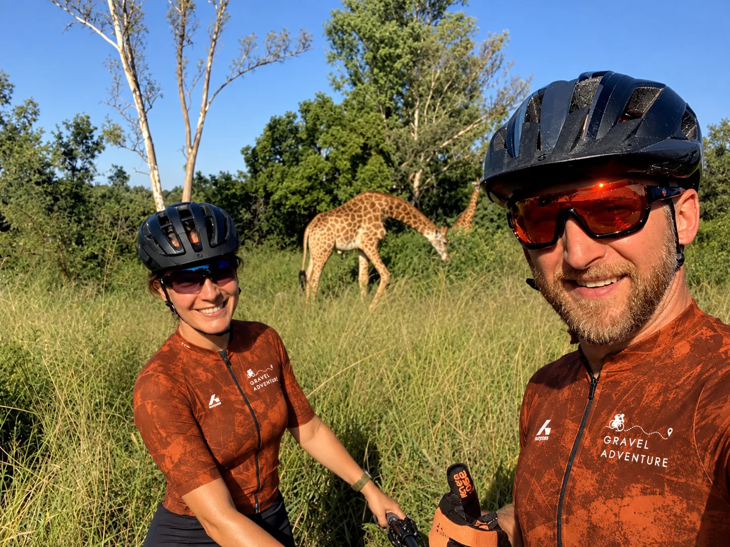 Tanya and Michael Sommer, founders of Gravel Adventure, on a gravel ride in South Africa with giraffes grazing behind