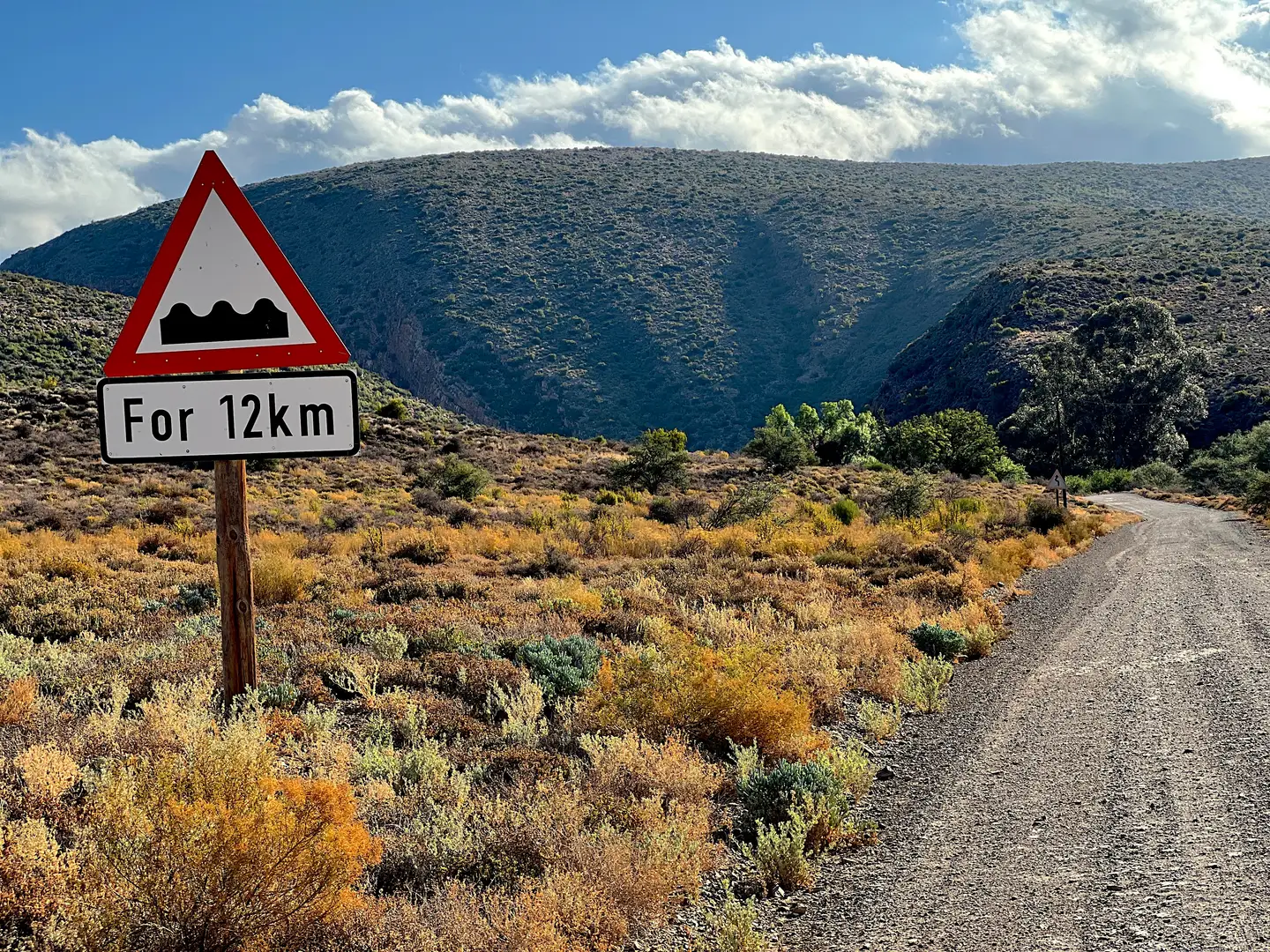 Rough-surface warning sign reading 'for 12 km' beside a gravel road through dry scrubland