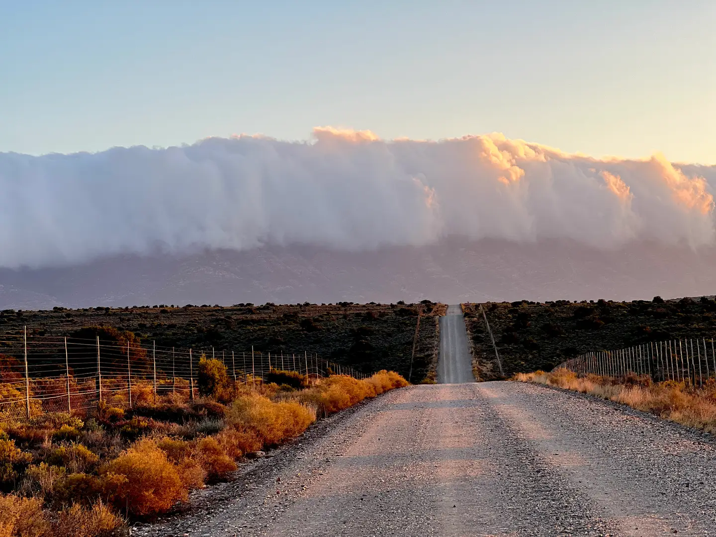 Gravel road running straight toward a wall of cloud over the mountains in golden light