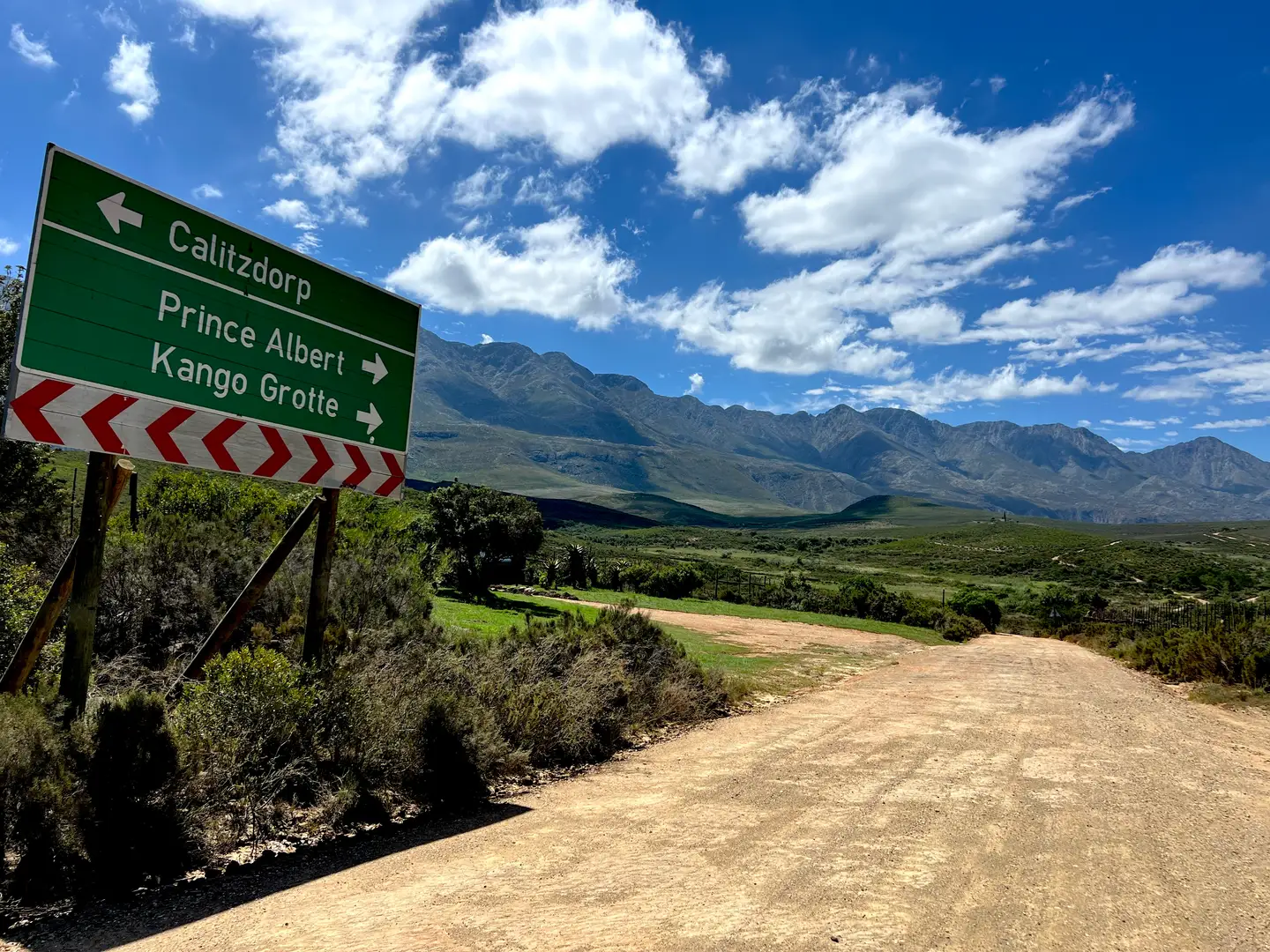 Gravel road below a mountain range, with a signpost pointing to Calitzdorp, Prince Albert and Kango Caves