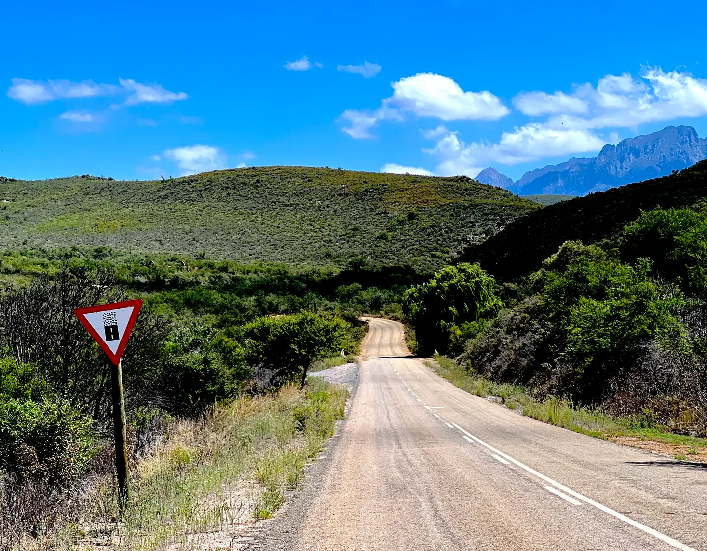 Tarmac road with a loose-surface warning sign, curving into green hills with mountains beyond