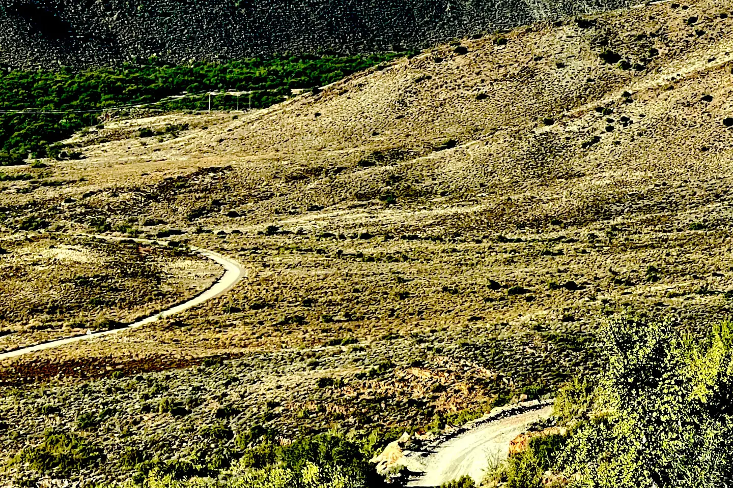 Gravel road winding across dry hills toward a green valley and distant mountains